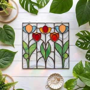 Op de afbeelding: Vierkant gebrandschilderd glaspaneel met een bloemenontwerp van rode, oranje en gele tulpen met groene stelen en bladeren. De witte achtergrond heeft een wervelend patroon, omlijst door donker metaal. Afmetingen: ongeveer 25 cm x 25 cm.
