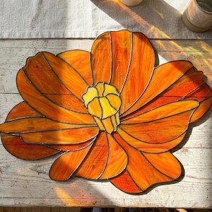May include: A stained glass flower in shades of orange and yellow. The flower has multiple petals and a detailed center. The glass is set in a dark frame, creating a striking contrast. The piece is likely a decorative item, possibly for a window or wall.