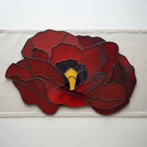 May include: A stained glass artwork depicting a large red poppy flower. The petals are various shades of red, with a black center and a yellow stamen. The artwork is set against a neutral background.