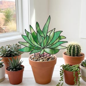 May include: A stained glass agave plant sculpture with variegated green glass panels and dark metal soldering. The sculpture is anchored in a terracotta pot filled with small, smooth, multi-coloured river stones, surrounded by various live potted succulents.