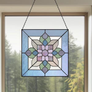May include: Stained glass window panel with a floral design. The square panel features a central flower in shades of purple, surrounded by blue, green, and neutral-toned glass. It hangs from a black chain, offering a decorative touch.