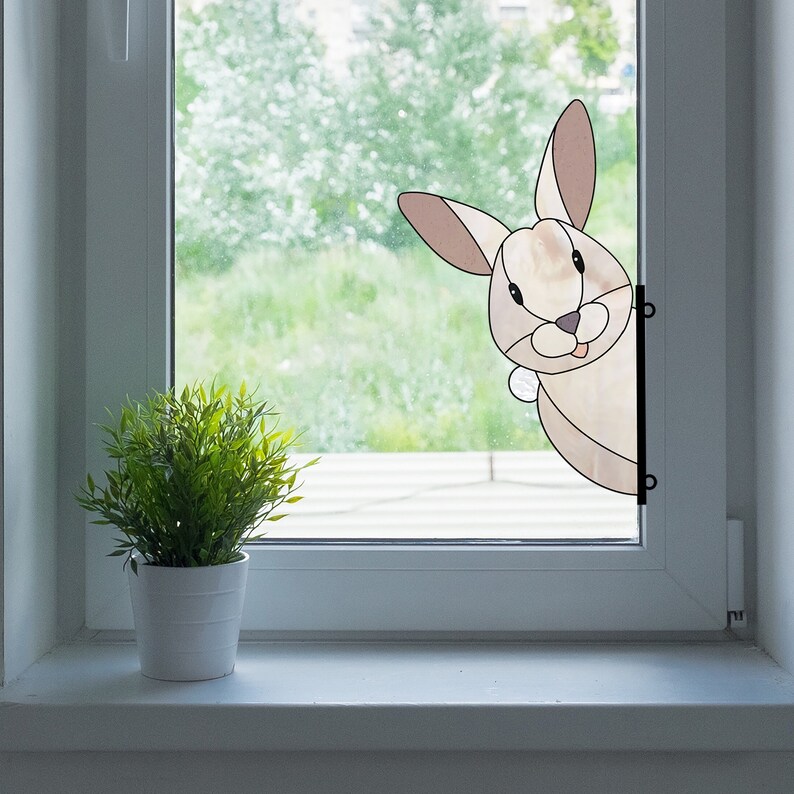 May include: A stained glass window cling of a brown rabbit peeking around the corner of a window frame. The rabbit has a white nose and a pink tongue sticking out.