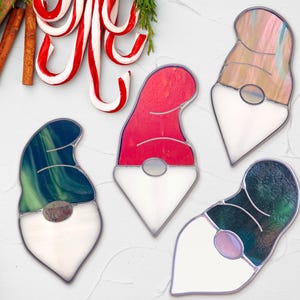 May include: Four stained glass gnome ornaments with white beards and different colored hats. The hats are red, green, blue, and pink.