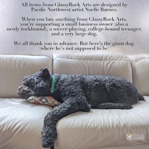 May include: A black dog with a green collar is lying on a white couch. The dog is sleeping and has its head resting on a pillow with a geometric pattern. The text on the wall behind the dog reads: "All items from GlassyRock Arts are designed by Pacific Northwest artist Noelle Barnes. When you buy anything from GlassyRock Arts, you're supporting a small business owner (also a nerdy rockhound), a soccer-playing, college-bound teenager, and a very large dog. We all thank you in advance. But here's the giant dog. (where he's not supposed to be)"