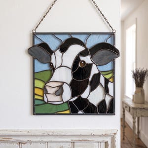 May include: A square stained glass panel featuring a black and white cow against a light blue sky and green pasture background. The piece is framed in dark metal and hangs from a metal chain, showcasing intricate glass segments and solder lines.