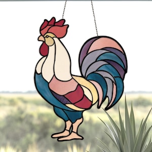 May include: A stained glass rooster suncatcher with a black outline. The rooster features a red comb, a cream-colored body, and a blue tail. The suncatcher hangs from a chain and is displayed in front of a blurred outdoor background.