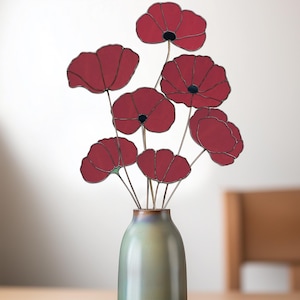 May include: A bouquet of eight red stained glass poppies in a light green vase. The poppies have black centers and are arranged in a vase with a metallic finish.