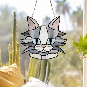 Puede incluir: Un atrapasoles de cristal con forma de cara de gato, con una combinación de colores gris y blanco con acentos rosas. La cara del gato está delineada en negro y cuelga de una cadena. Los ojos del gato son azules y la nariz es rosa.