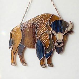 May include: Stained glass bison wall hanging. The artwork features a bison in shades of brown, blue, and tan, with copper-colored lines defining the shapes. The piece is suspended by a delicate chain, adding a touch of elegance to the rustic design.