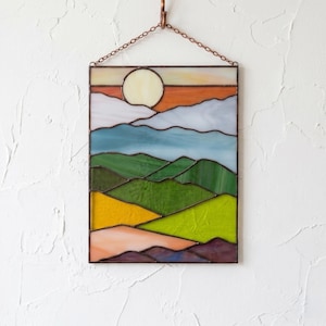May include: A rectangular stained glass panel featuring a landscape scene with a pale yellow sun, orange sky, light blue clouds, and layered green hills. The bottom section displays yellow, peach, and purple glass segments, all framed in copper.