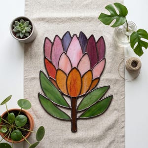 May include: A stained glass suncatcher shaped like a protea flower, featuring petals in shades of pink, purple, and orange, with green glass leaves. The piece is constructed with dark metal solder lines outlining each individual glass segment.