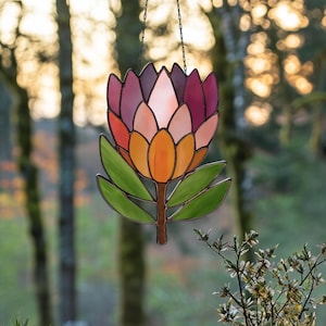 Puede incluir: Un atrapasoles de vidrio coloreado con forma de flor, con una vibrante paleta de colores naranja, rosa y morado. La flor está suspendida por una cadena, con hojas verdes y un tallo marrón. El fondo es un bosque borroso.