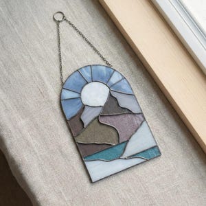 May include: Stained glass window panel depicting a mountain landscape with a sun. The panel features a variety of colors, including blue, white, gray, and purple. It is suspended by a metal chain and ring.