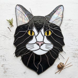 May include: A stained glass cat head suncatcher featuring black and white glass pieces, bright yellow eyes, a soft pink nose, and thin metal wire whiskers. The ears are crafted from iridescent glass that reflects light in various colors.
