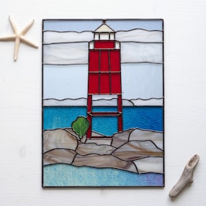 May include: A rectangular stained glass panel depicting a red lighthouse standing on rocky terrain against a blue sea and sky. The scene includes a small green bush near the base of the lighthouse, with a white starfish and a piece of driftwood nearby.
