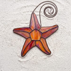 May include: A stained glass starfish ornament featuring a vibrant blend of orange, yellow, and purple hues. The five-pointed star is constructed with dark metal solder lines and includes a matching metal chain for hanging, creating a decorative piece.