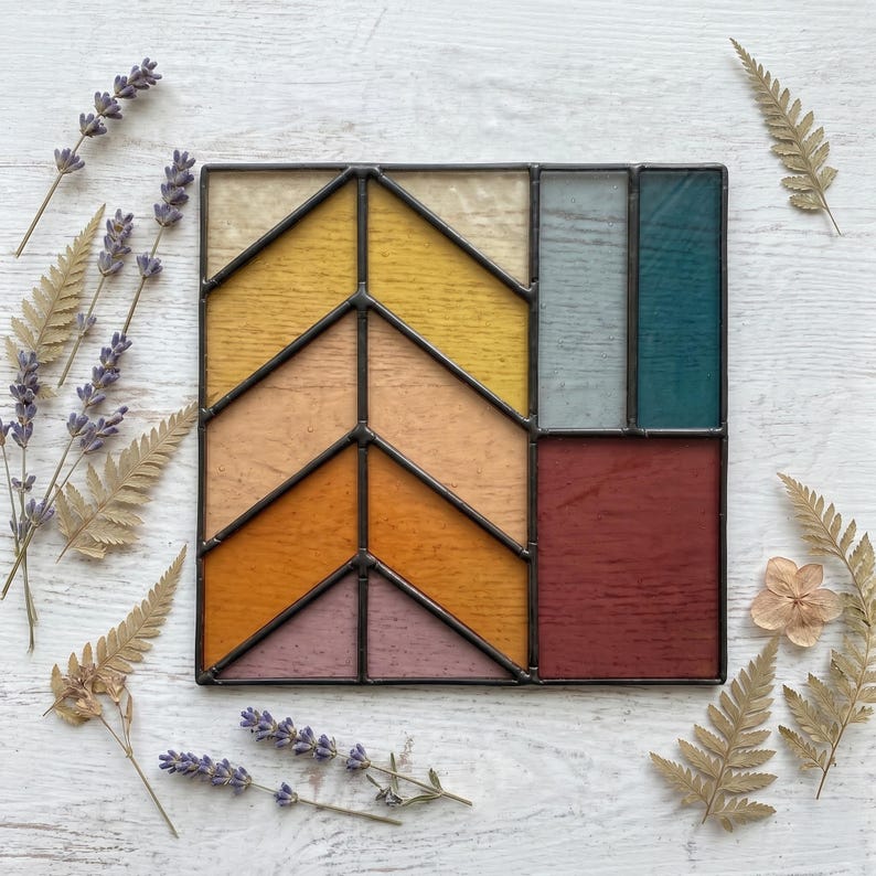 May include: A square stained glass piece with a geometric design. The design features a series of angled shapes in shades of yellow, orange, pink, teal, and burgundy, framed by black metal. Dried lavender and ferns surround the glass.