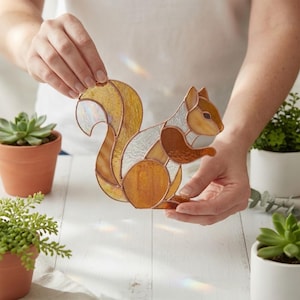 Könnte beinhalten: Glasmalerei-Eichhörnchen-Figur mit einem mosaikartigen Design. Das Eichhörnchen hat Farbtöne von Bernstein, Braun und klarem Glas, mit einem großen, buschigen Schwanz. Die Figur wird vor einer weißen Oberfläche gehalten.
