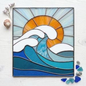 Könnte beinhalten: Glasfenster mit einer stilisierten Ozeanwelle und untergehender Sonne. Das Kunstwerk verwendet Blau-, Weiß- und Orangetöne, gerahmt in dunklem Metall. Die Sonne ist orange und die Welle hat weißen Schaum. Das Panel ist quadratisch.