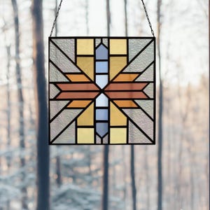 Puede incluir: Panel de ventana cuadrado de vidrieras con un diseño geométrico. El panel presenta un borde negro y un patrón de piezas de vidrio naranja, amarillo, azul y transparente. Cuelga de una cadena sobre un fondo borroso de árboles.