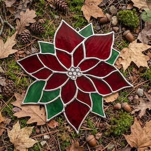 May include: A stained glass poinsettia ornament featuring deep red petals and vibrant green leaves, constructed with individual glass segments joined by silver-toned metal solder lines, centred with a small cluster of white glass circles.