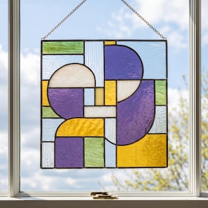 Peut inclure: Panneau de vitrail rectangulaire avec un motif géométrique. Le panneau présente des formes de différentes couleurs : violet, jaune, vert, blanc et bleu. Il est suspendu par une chaîne devant une fenêtre.