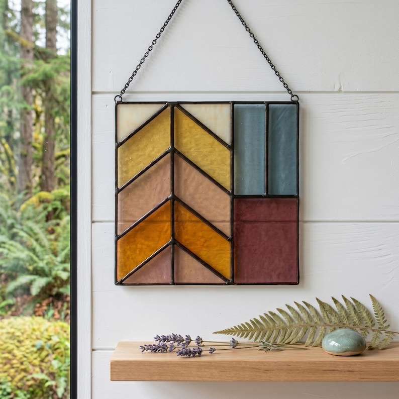 May include: A geometric stained glass panel with a black metal frame. The design features a chevron pattern in shades of yellow, orange, and pink, with blue and burgundy rectangular sections. It hangs from a chain.