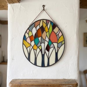 May include: A circular stained glass panel featuring an abstract tree motif with a dark metal frame. The design consists of numerous multi-colored glass segments in shades of orange, yellow, blue, green, and white, suspended by a metal chain.