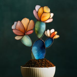 Puede incluir: Una escultura de cactus de vidrieras con flores coloridas en tonos naranja, rosa y blanco. El cactus es azul y verde, colocado en una maceta de cerámica blanca llena de tierra. El fondo es verde oscuro.