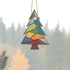 May include: A stained glass Christmas tree ornament. The tree is composed of various colored glass pieces, including red, blue, green, yellow, and white, framed by copper. It hangs from a chain against a blurred forest background.