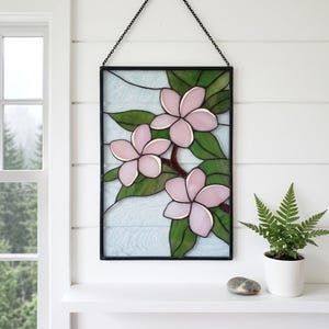 May include: Stained glass panel featuring three pink flowers with green leaves on a brown branch. The artwork is framed in black and hangs from a chain. The background is a light blue color.