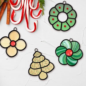 Puede incluir: Una colección de adornos navideños de vidrieras en varias formas: una corona, una flor, una flor de cuatro pétalos y un árbol de Navidad. Los adornos tienen contornos negros y están decorados con detalles rojos y verdes. Bastones de caramelo y palitos de canela en el fondo.