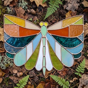 May include: A stained glass butterfly with copper edging. The wings feature a mosaic of colors including yellow, orange, teal, blue, green, and white. The butterfly is resting on a bed of leaves, moss, and acorns.