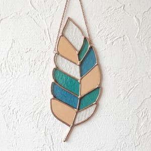 Pode incluir: Um ornamento de vitral em forma de folha com uma moldura de metal acobreado e uma corrente delicada para pendurar. A folha é composta por segmentos de vidro individuais em tons alternados de bege suave, verde-azulado e azul profundo.