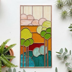 Op de afbeelding: Rechthoekig glas-in-loodpaneel met een landschap van bomen en wolken. Het kunstwerk gebruikt groen, geel, oranje, rood en blauw glas, omlijst met koper. Het paneel toont een gestileerde buitenscène.