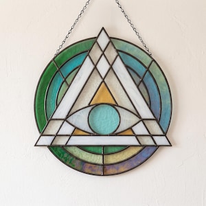 Pode incluir: Um apanhador de sol circular em vitral com um motivo central de triângulo e olho. A peça é construída com segmentos de vidro branco, azul-petróleo, verde e amarelo unidos por linhas de solda de metal escuro, suspensa por uma fina corrente preta.