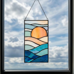 Puede incluir: Panel de vidrieras que representa una puesta de sol sobre las olas del océano. La obra de arte presenta un sol naranja, olas azules y turquesas, y una cadena para colgar. El panel es rectangular y está enmarcado en negro.