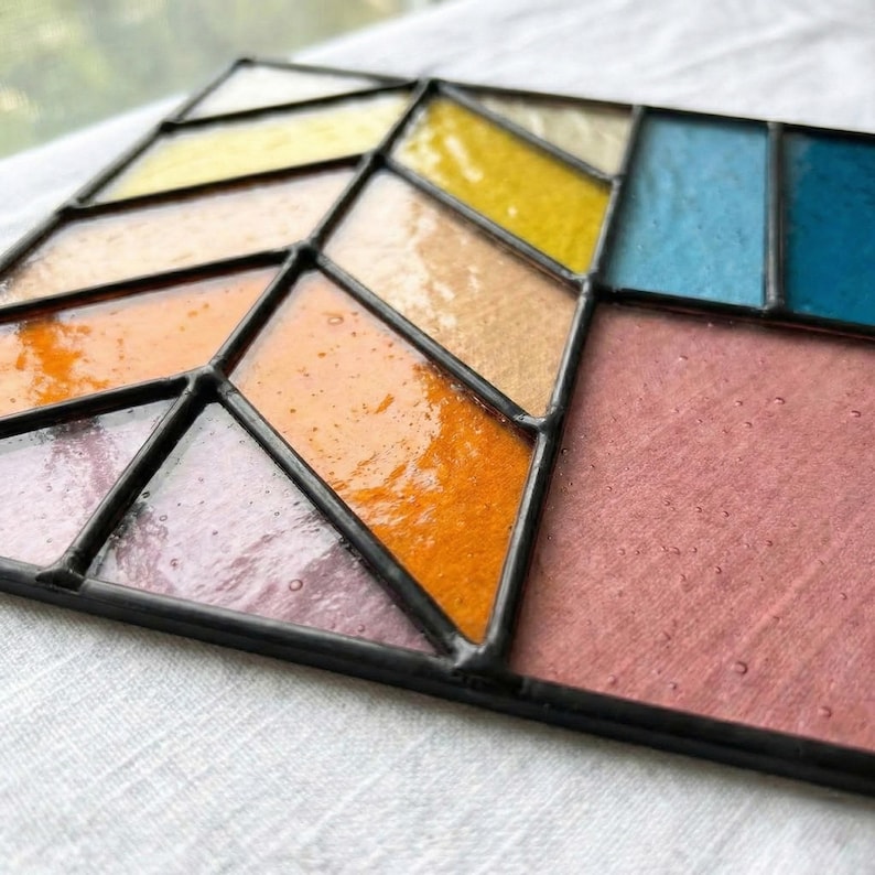 May include: A stained glass panel with a geometric design. The panel features various colors, including yellow, orange, pink, blue, and clear glass, all framed by black lines. The glass has a glossy finish.