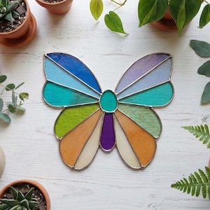 May include: A colorful stained glass butterfly with a silver metal outline. The wings feature sections of blue, teal, green, orange, and purple glass. The butterfly is surrounded by plants and small terracotta pots.