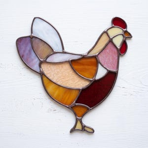 Könnte beinhalten: Ein Sonnenfänger aus Buntglas in Form eines Hahns, zusammengesetzt aus verschiedenen farbigen Glasstücken in Weiß, Violett, Bernstein, Orange und Dunkelrot, die durch dunkle Metalllötlinien zu einer stehenden Vogelgestalt verbunden sind.