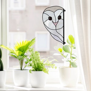 May include: A stained glass window hanging featuring a white owl design. The owl has large black eyes and a pink beak. The owl is peeking out from behind a white curtain.