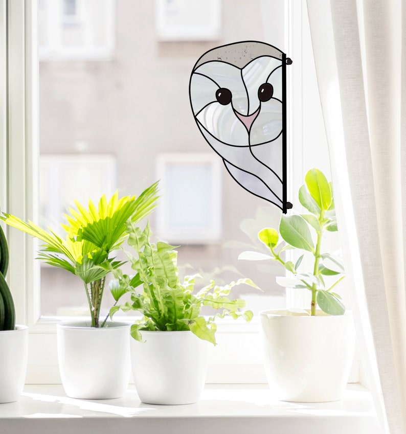 May include: A stained glass window hanging featuring a white owl design. The owl has large black eyes and a pink beak. The owl is peeking out from behind a white curtain.