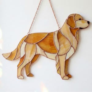 Op de afbeelding: Gebrandschilderde hondensilhouet in tinten amber, crème en goud, met koperkleurige metalen contouren. De hond hangt aan een delicate ketting, perfect voor raam- of wanddecoratie. Het kunstwerk toont een Golden Retriever.