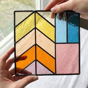 May include: A stained glass panel with a black metal frame. The panel features a geometric design with various colors, including yellow, orange, pink, and blue. The panel is held by a person's hands.