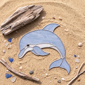 May include: A stained glass dolphin suncatcher featuring marbled light blue and white glass panels held together by thin metal solder lines. The dolphin has a single black glass bead for an eye and is depicted in a curved, leaping pose.