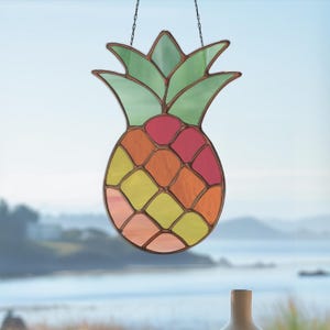 May include: A stained glass pineapple suncatcher with a copper-colored frame. The pineapple features a green crown and a body of colorful glass pieces in shades of pink, orange, yellow, and peach. It hangs from a chain, perfect for a window.