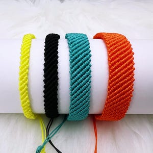 Puede incluir: Cuatro pulseras de amistad trenzadas en amarillo, negro, azul verdoso y naranja. Las pulseras están hechas con un patrón tejido y tienen un cierre anudado.