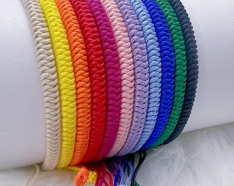 Può includere: Una collezione di braccialetti colorati intrecciati in un arcobaleno di tonalità, tra cui beige, giallo, arancione, rosso, rosa, lavanda, azzurro, blu scuro, verde e nero. Ogni braccialetto ha una chiusura regolabile con coulisse.