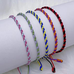 Puede incluir: Cinco pulseras de amistad trenzadas de colores. Las pulseras son en tonos rosa, verde, azul, amarillo, naranja y rojo. Cada pulsera tiene cuerdas ajustables para el tamaño. Las pulseras se muestran sobre una superficie blanca.