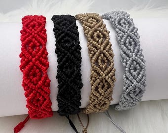 Può includere: Quattro braccialetti in macramè rosso, nero, beige e grigio. Ogni braccialetto presenta un motivo a diamante e lacci regolabili. I braccialetti sono esposti su una superficie bianca con uno sfondo morbido e piumato.